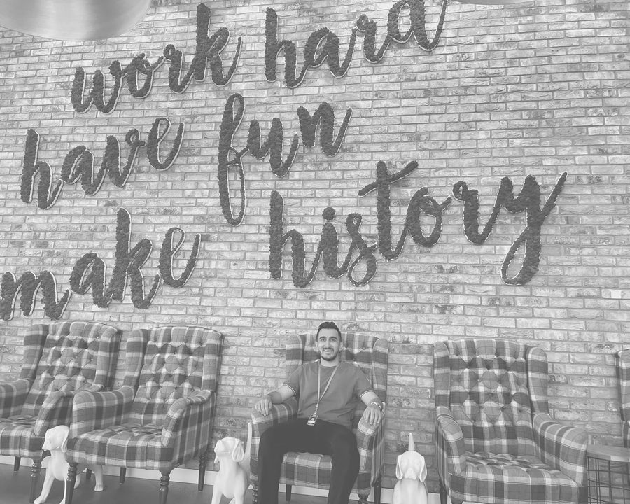 Salman sitting in an armchair against a brick office wall lettered work hard, have fun, make history.