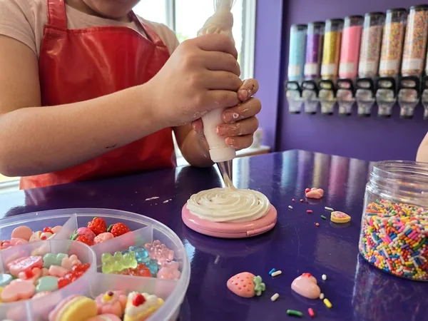 Decoden close-up: two hands piping white cream onto a pink compact, a tray of charms in front and the dispenser wall behind.