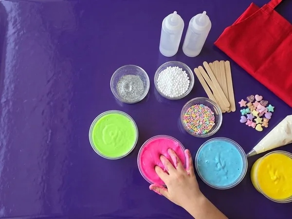 A workshop station from above: pots of coloured slime, glitter, beads and lolly sticks on a glossy purple table, a red apron, and one hand pressing into pink slime.